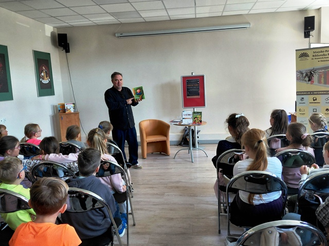 Pisarz stoi przed dziećmi i pokazuje swoją książkę „Kacperiada”. Grupa dzieci siedzi na krzesłach.