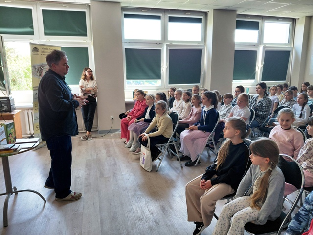Pisarz stoi przed grupą siedzących na krzesłach dziećmi.