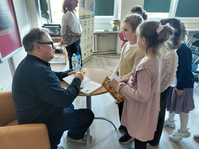 Autor siedzi przed stolikiem i podpisuje książkę.. Przed nim stoją dziewczynki.